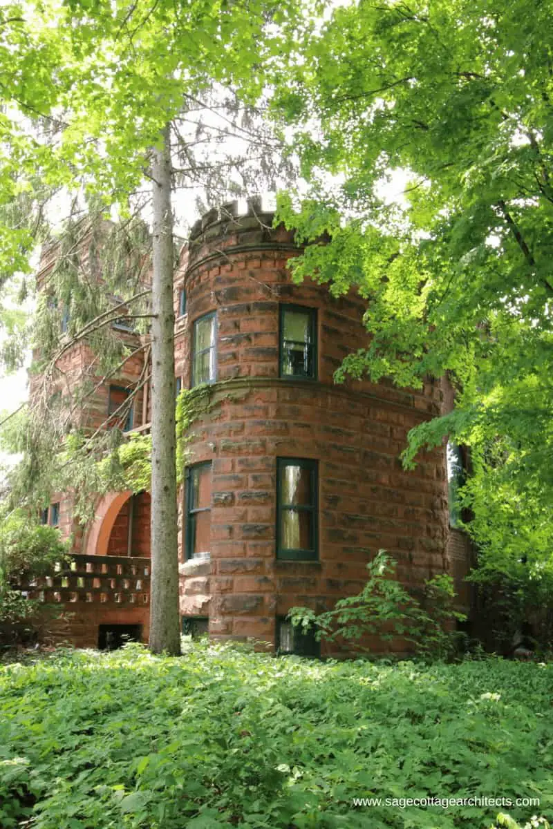 Side view of a typical Richardsonian Romanesque home - red ashlar walls with a round turret and lots of foliage.