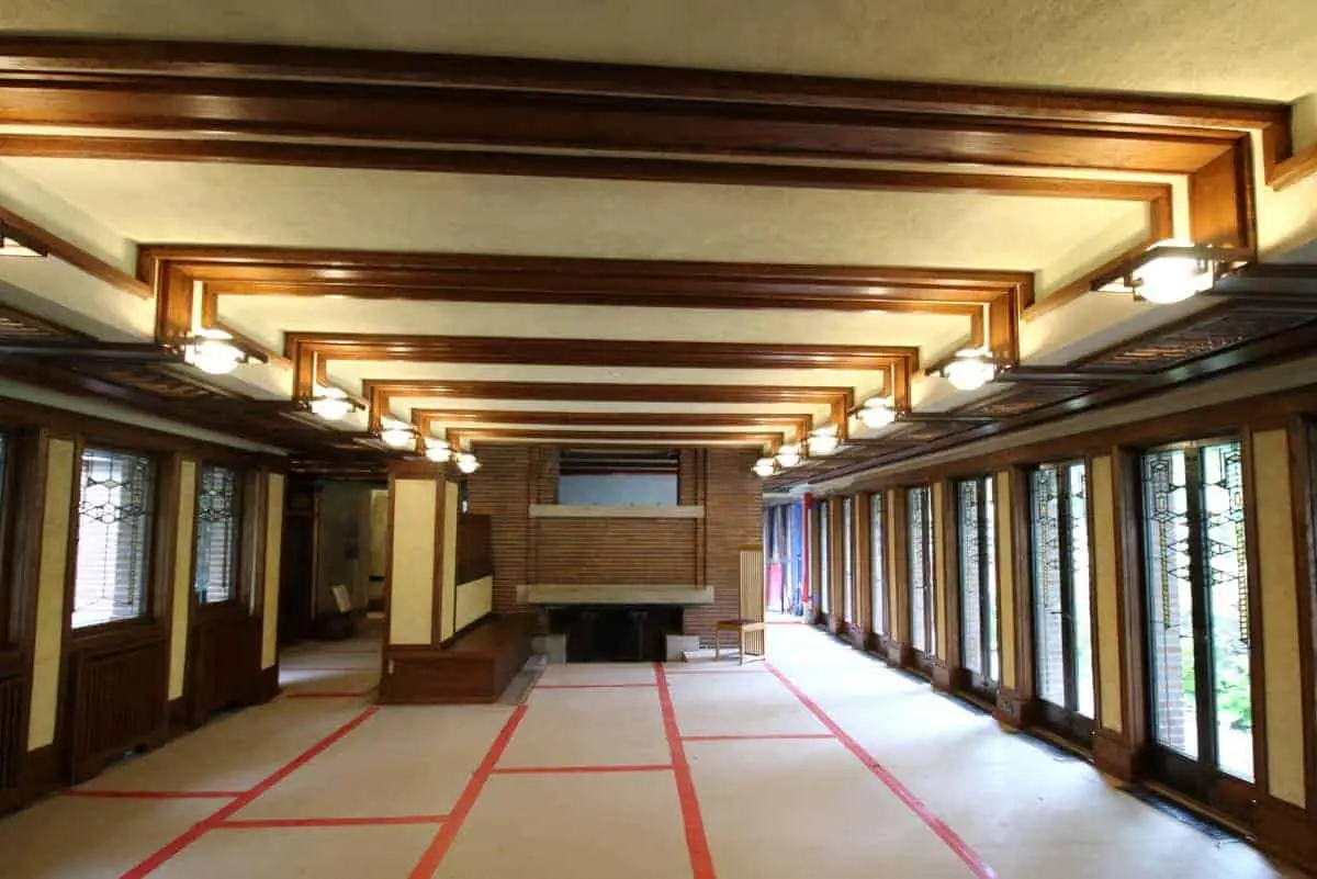 Prairie style home interior with oak ceiling beams, cream ceiling, windows along one wall and a central brick fireplace.