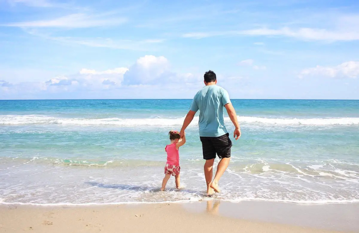 Photo of a dad and young daughter walking along the ocean's edge on a beach vacation. 