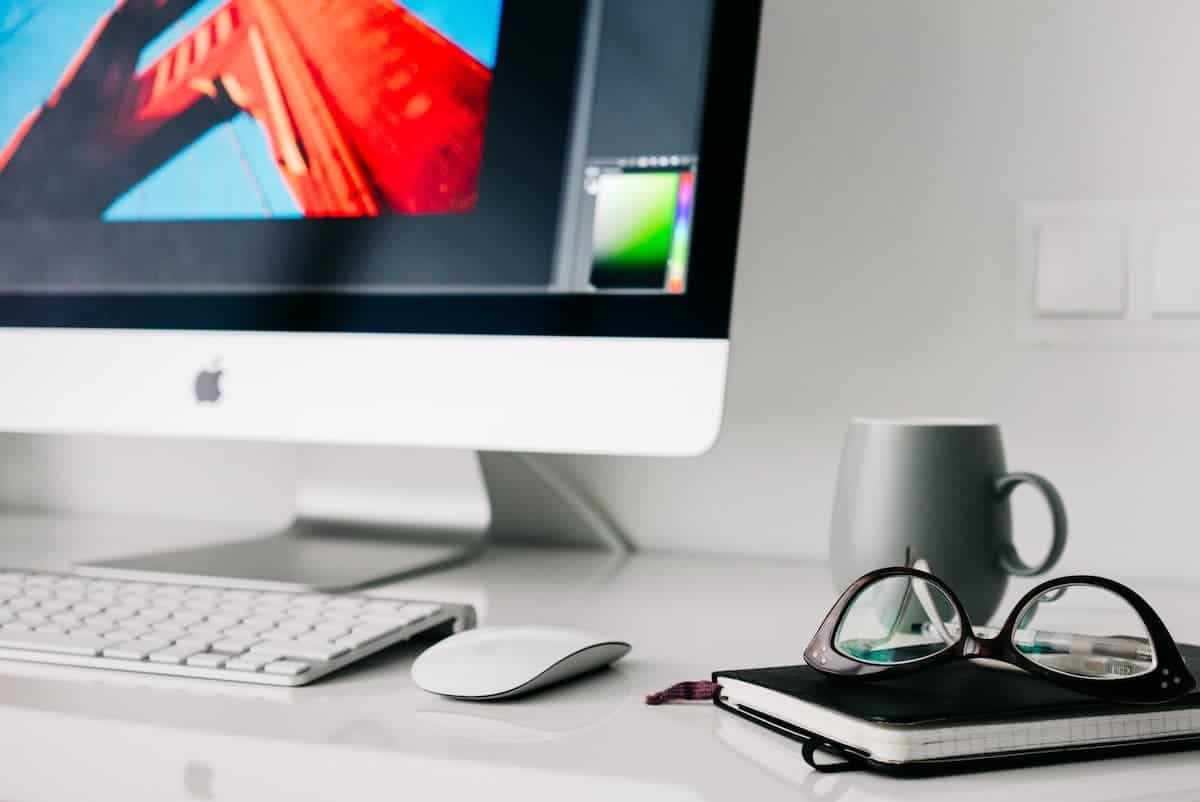 Photo of a home office with Apple computer, white desktop, mug, glasses and a notebook.