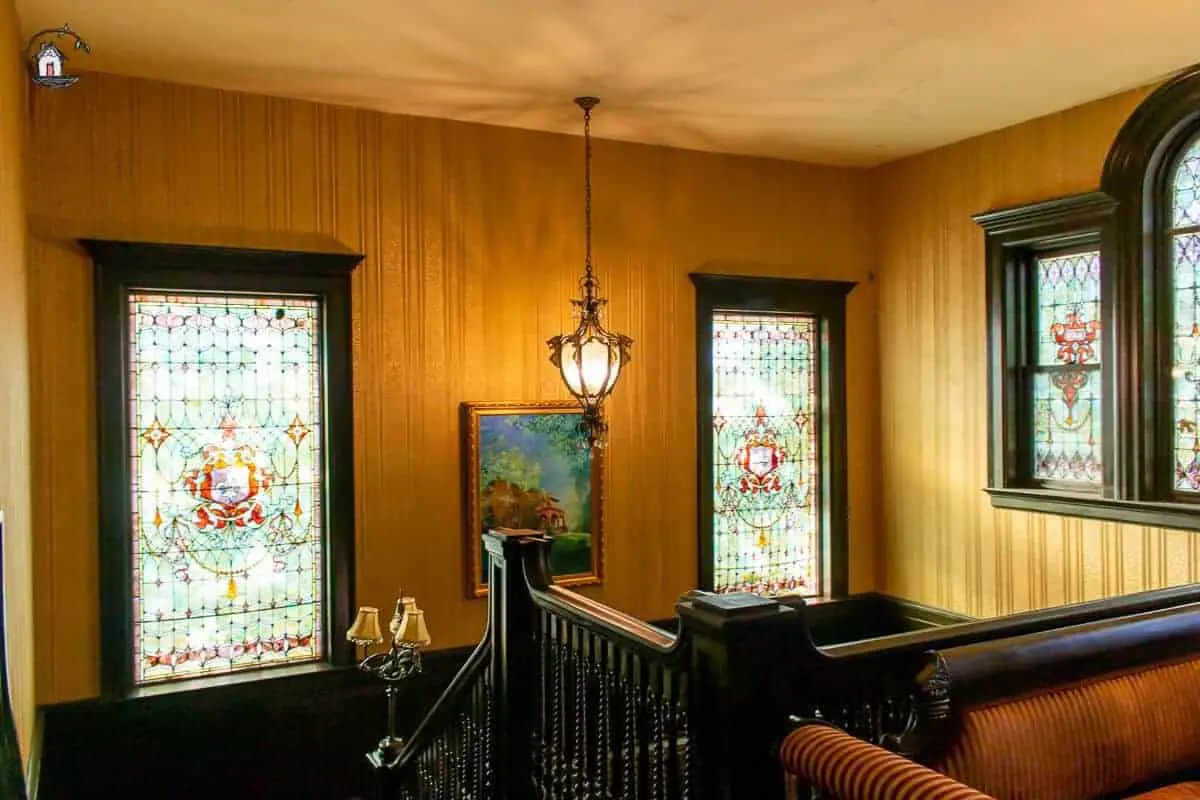 Photo of large stained glass windows, dark wooden handrails and hanging light in the stairway of the Vrooman Mansion. 