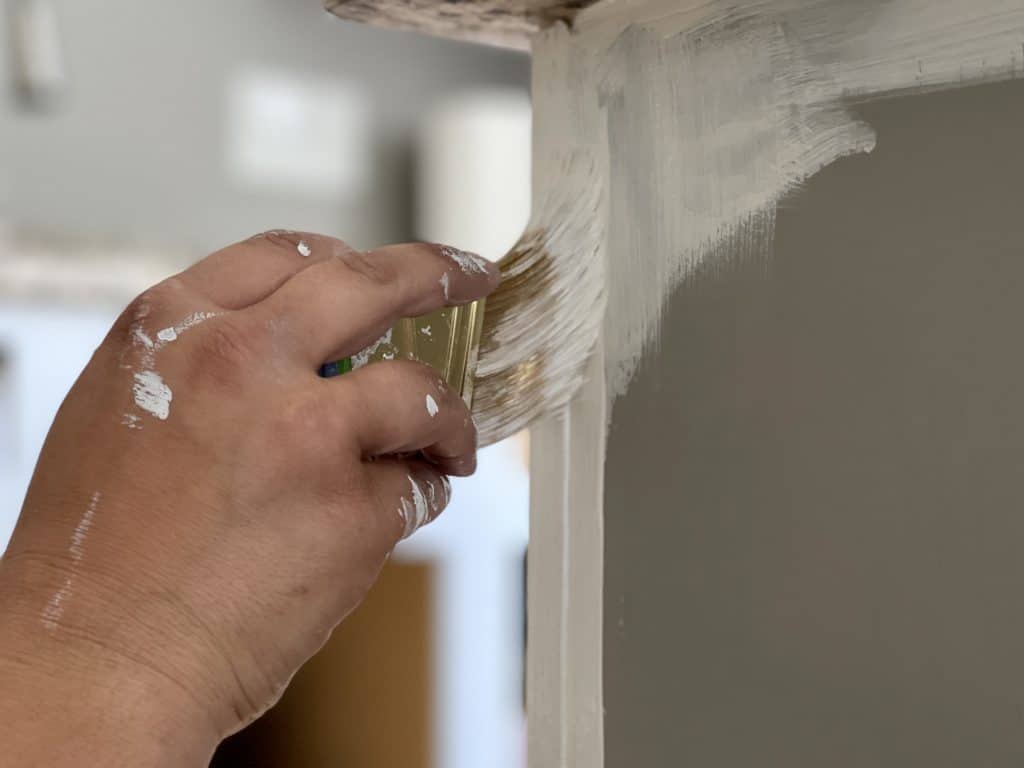 hand with paintbrush brushing white paint on cabinet body